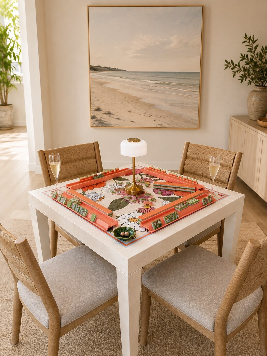 A mahjong table set for four in a sunlit dining room, with coral racks, a floral mat and champagne poured