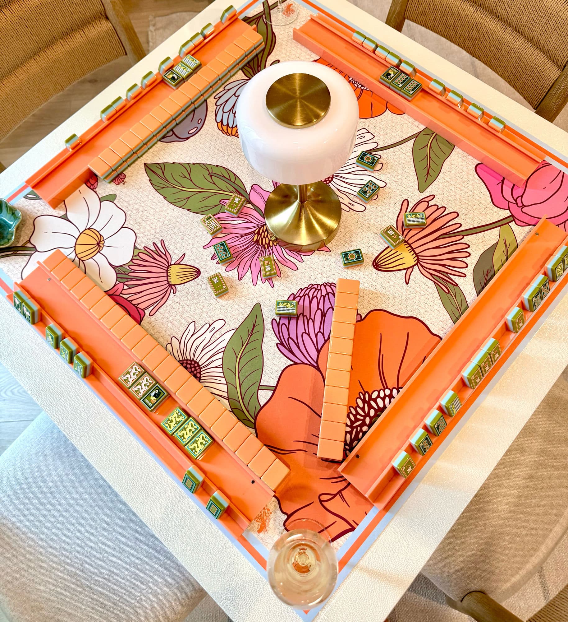 A coral mahjong set laid out on a poppy print mat, with a brass lamp and a glass of wine on the dining table