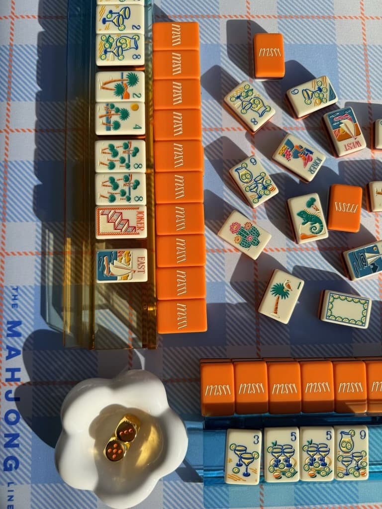 Orange and cream mahjong tiles with palm and flamingo motifs, laid out in sunlight on a blue plaid mat