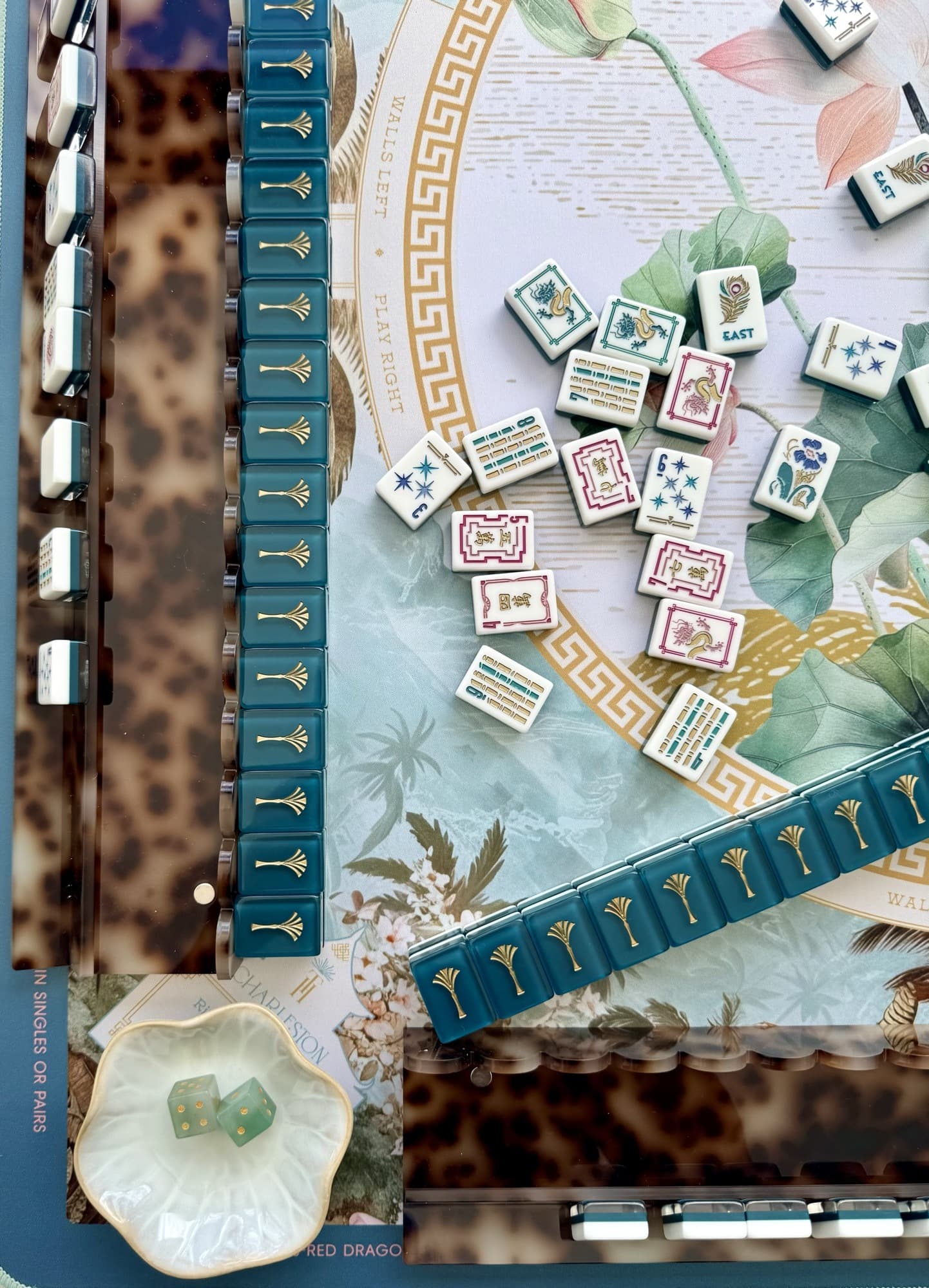 Teal and gold mahjong tiles in tortoiseshell racks on a lotus-patterned mat, with dice in a shell dish