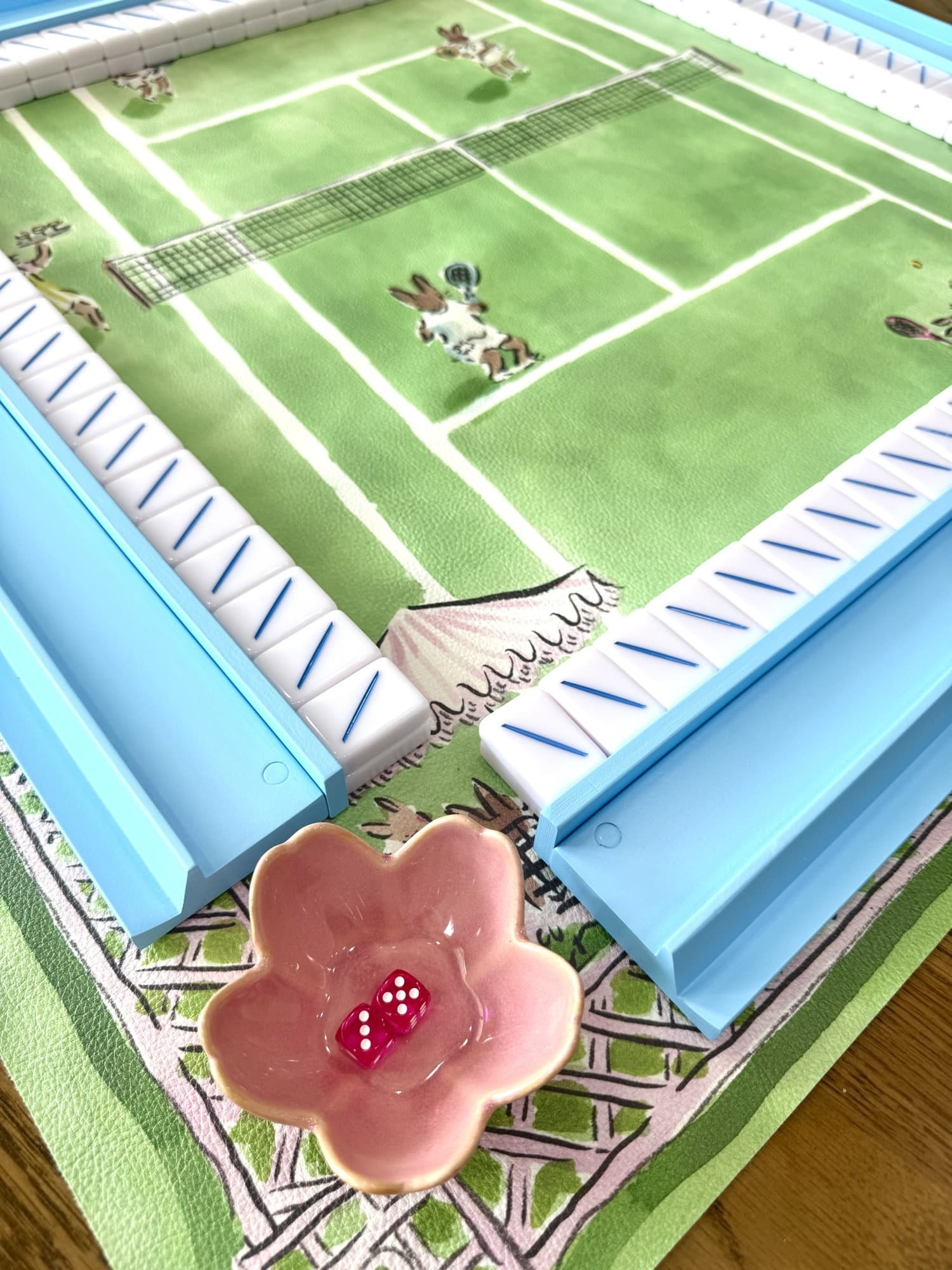 A mahjong table mid-game on a tennis court mat, with pale blue racks and pink dice in a flower dish